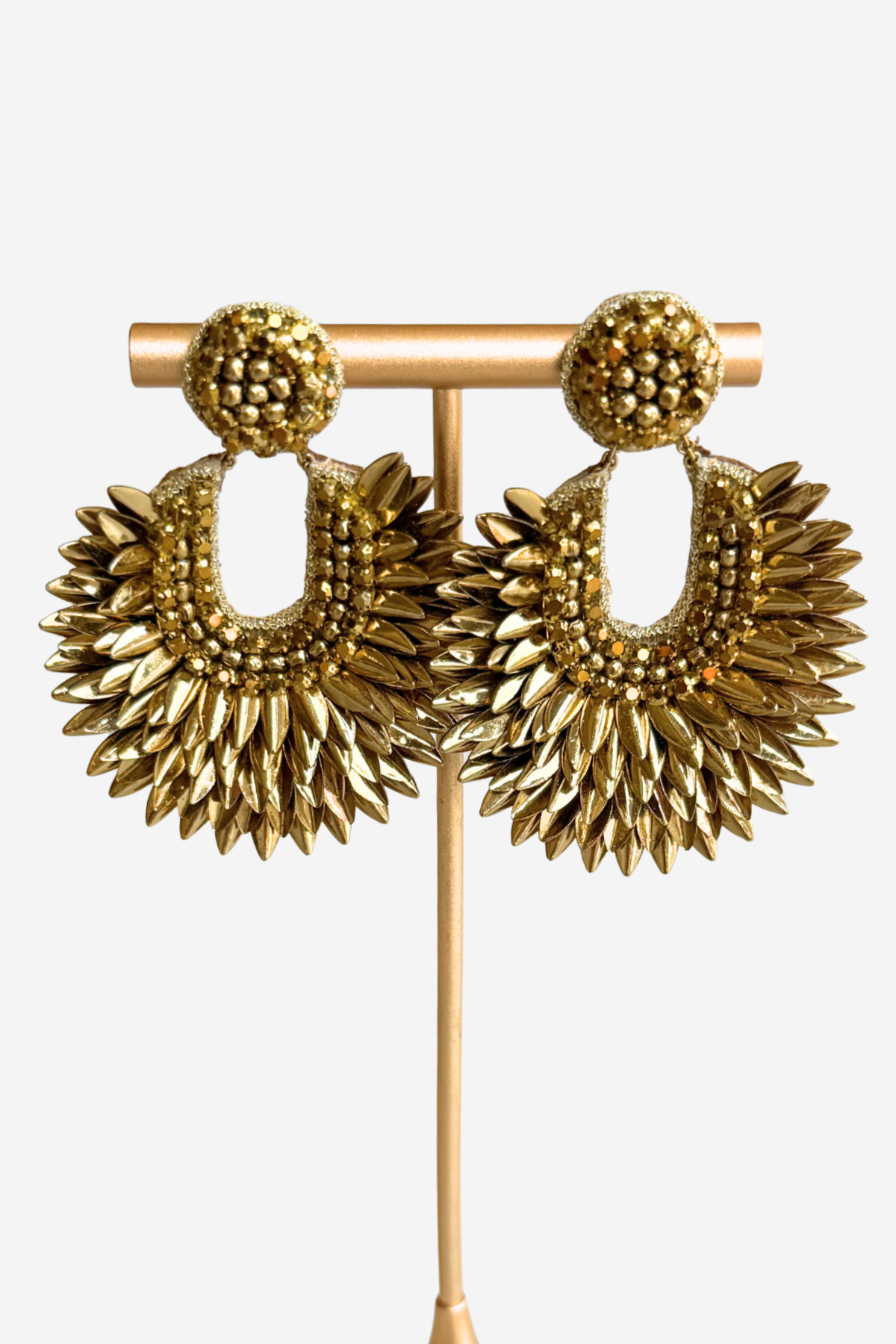 A pair of chunky gold-tone drop earrings displayed on a matching gold stand against a light gray background. The earrings feature textured spherical studs and large, sunburst-shaped dangles with layered beaded and metallic details.