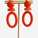 Bright orange beaded statement earrings displayed on a gold-tone stand against a light gray background.