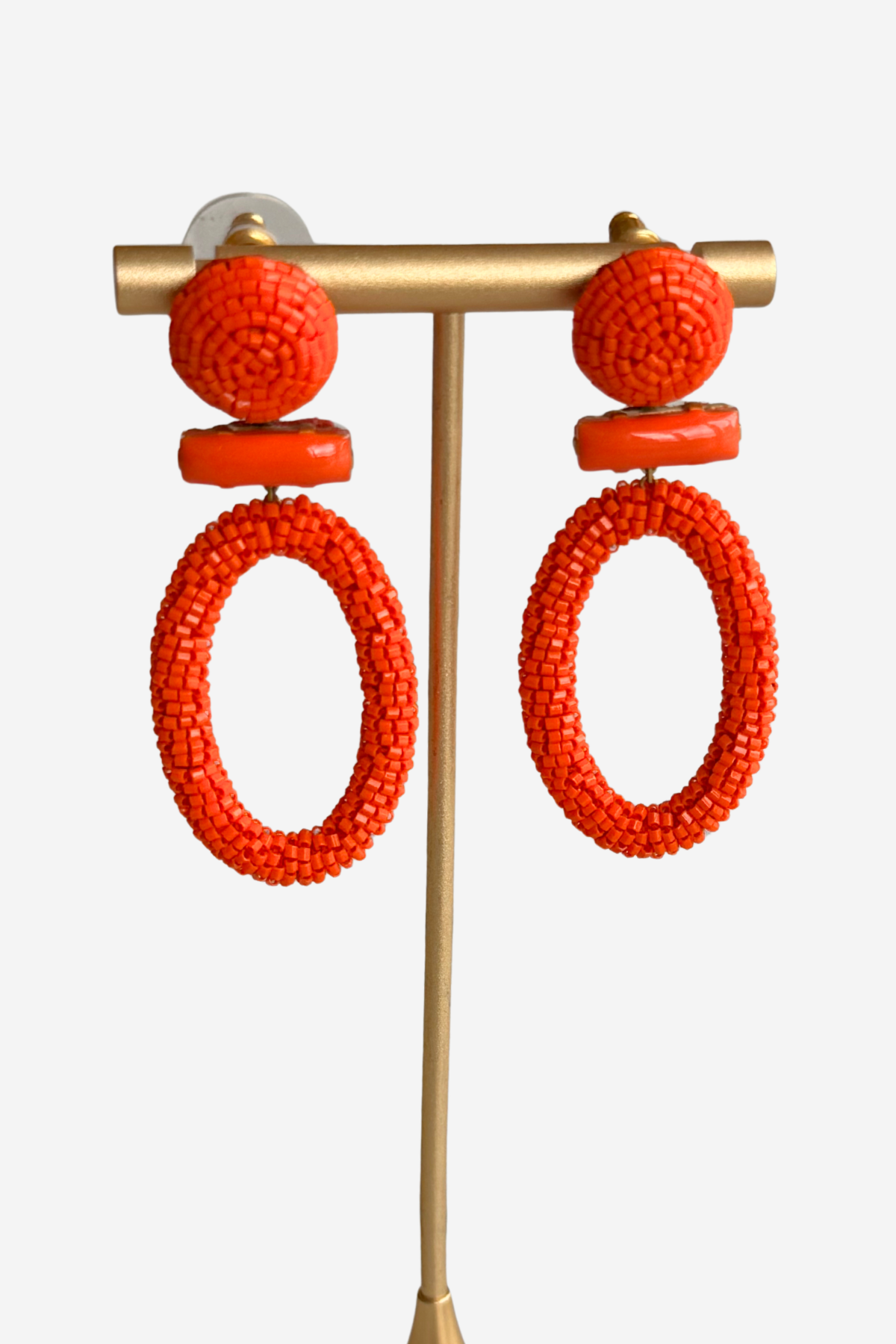 Bright orange beaded statement earrings displayed on a gold-tone stand against a light gray background.