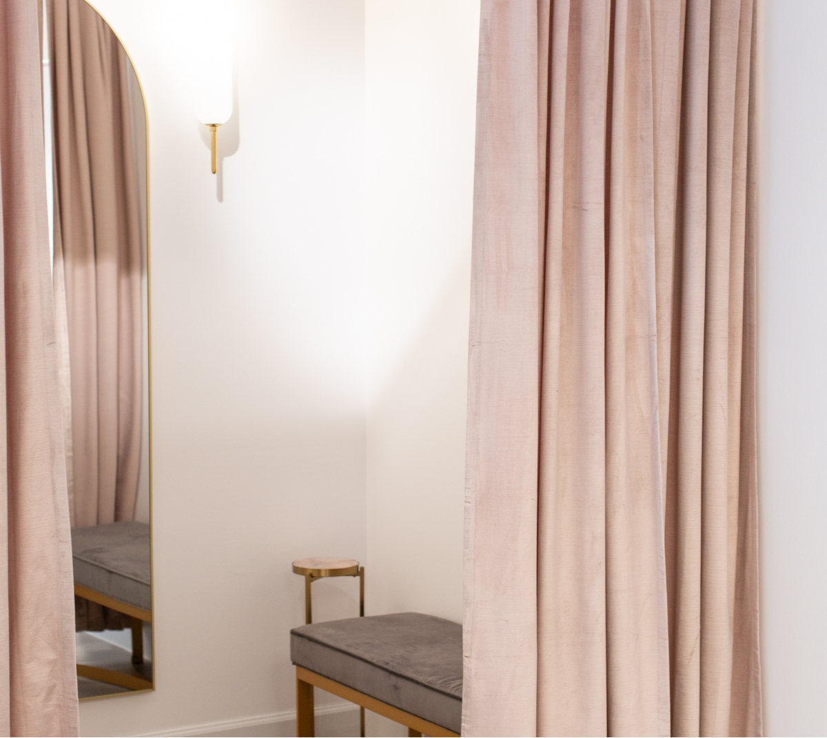 A pair of dusty blush pink velvet curtains hanging in a fitting room, with a mirror reflecting a gray padded bench with gold legs.