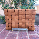 Brown woven clutch bag on a brick surface with blurred greenery in the background
