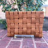 Brown woven clutch bag on a brick surface with blurred greenery in the background