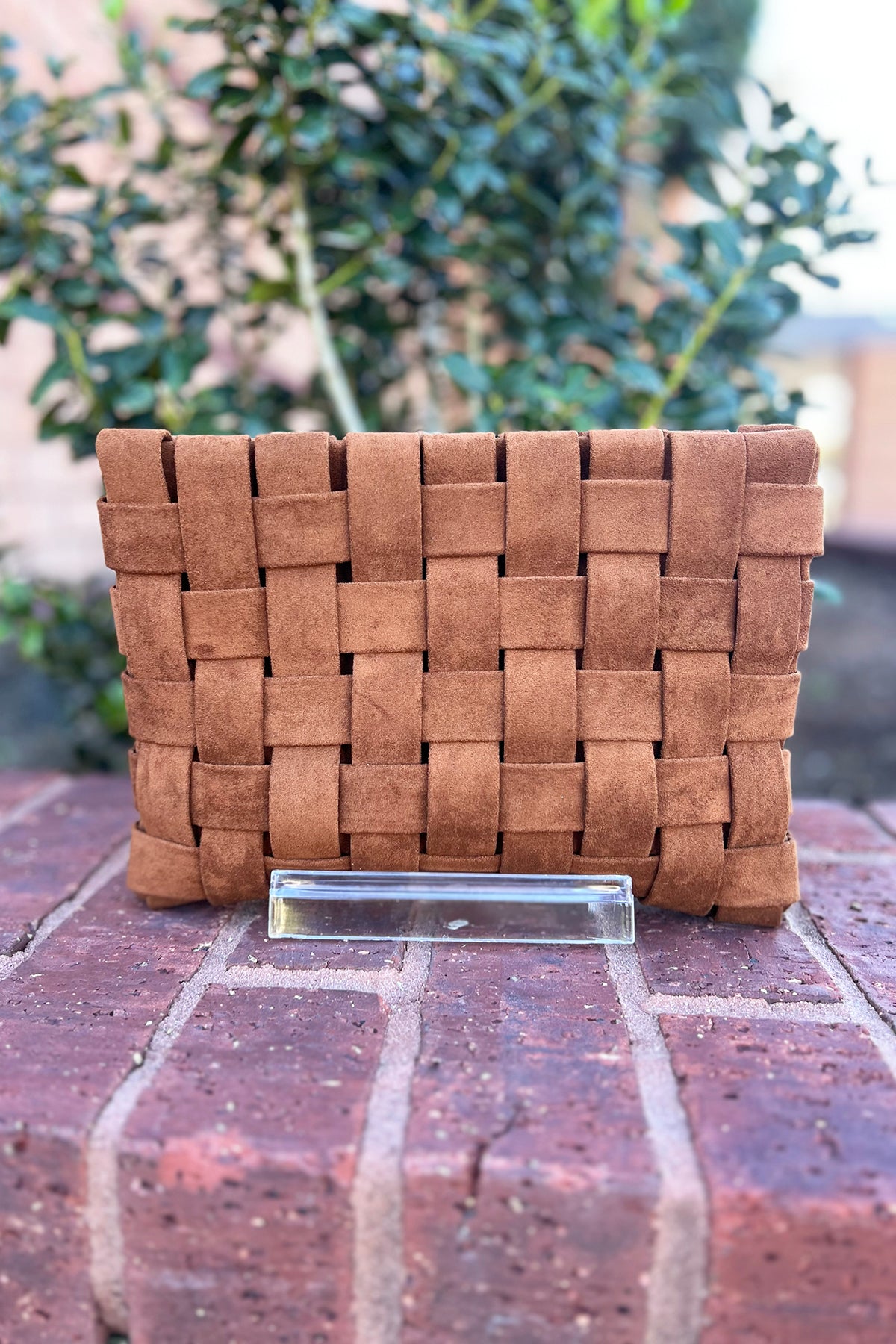 Brown woven clutch bag on a brick surface with blurred greenery in the background