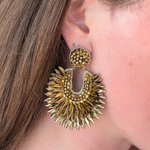 A close-up of a person's ear wearing a large, textured gold statement earring with a circular beaded top and a sunburst-shaped lower dangle.