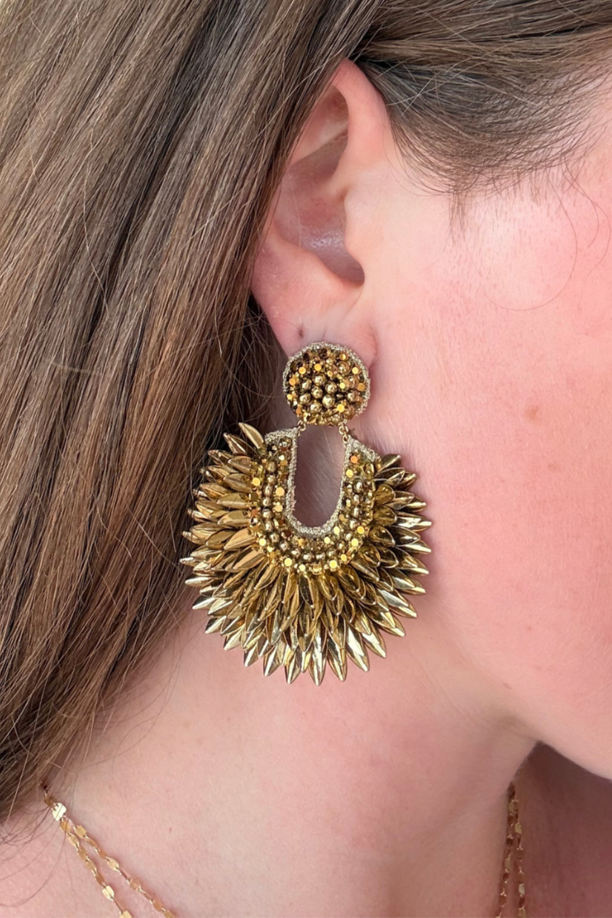 A close-up of a person's ear wearing a large, textured gold statement earring with a circular beaded top and a sunburst-shaped lower dangle.
