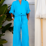 Person wearing a bright blue jumpsuit standing outdoors with plants and a white wall in the background.