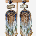 A pair of statement dangle earrings displayed on a gold-tone stand against a light gray background. The earrings feature a round blue and gold beaded top, a clear faceted stone, and long cascading fringe made of gold, blue, and light blue beads.