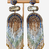 A pair of statement dangle earrings displayed on a gold-tone stand against a light gray background. The earrings feature a round blue and gold beaded top, a clear faceted stone, and long cascading fringe made of gold, blue, and light blue beads.