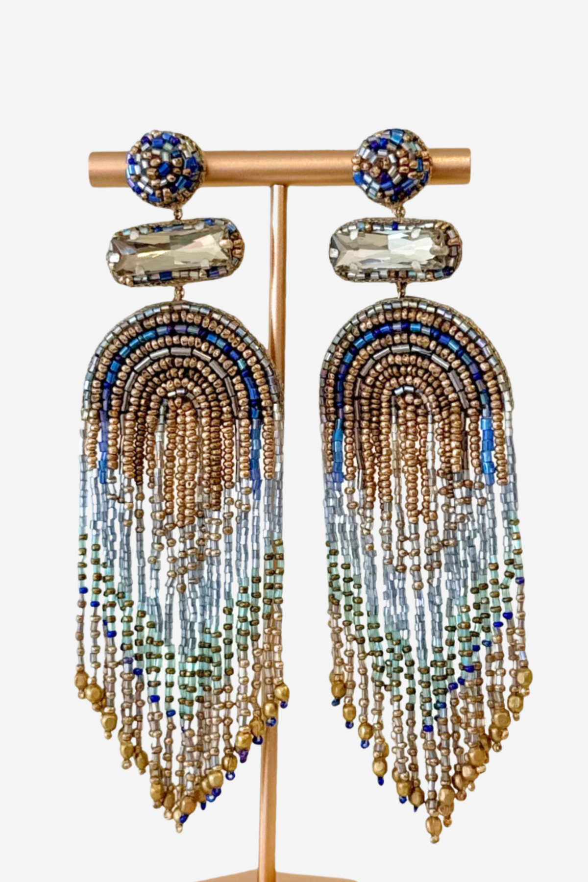 A pair of statement dangle earrings displayed on a gold-tone stand against a light gray background. The earrings feature a round blue and gold beaded top, a clear faceted stone, and long cascading fringe made of gold, blue, and light blue beads.