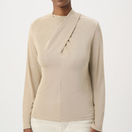 A woman modeling a beige long-sleeve wrap-style top with a mock neck and asymmetrical draped front detail, paired with light cream pants, against a plain light gray studio background.