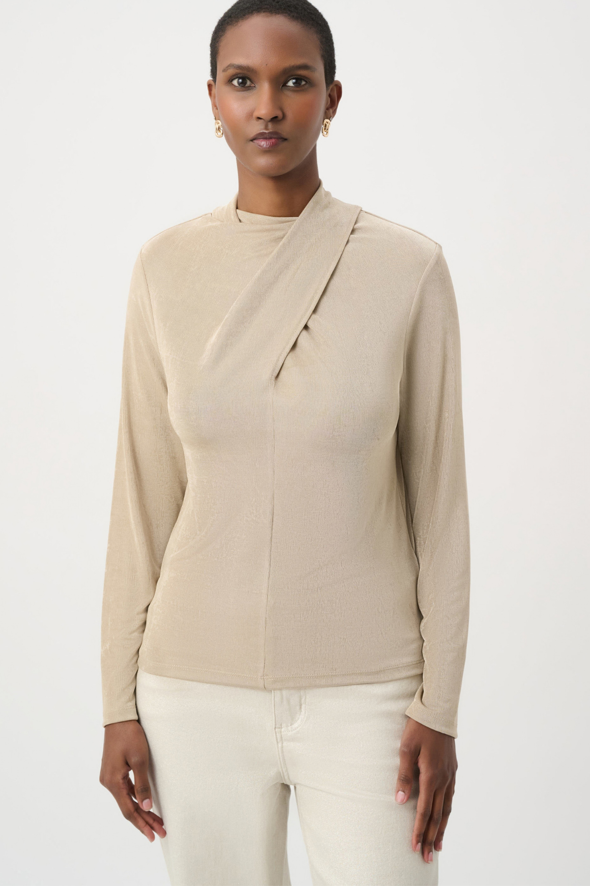 A woman modeling a beige long-sleeve wrap-style top with a mock neck and asymmetrical draped front detail, paired with light cream pants, against a plain light gray studio background.