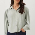 A woman wearing a light sage green long-sleeve button-up blouse with gathered shoulders and dark blue jeans, standing against a plain light gray studio background.