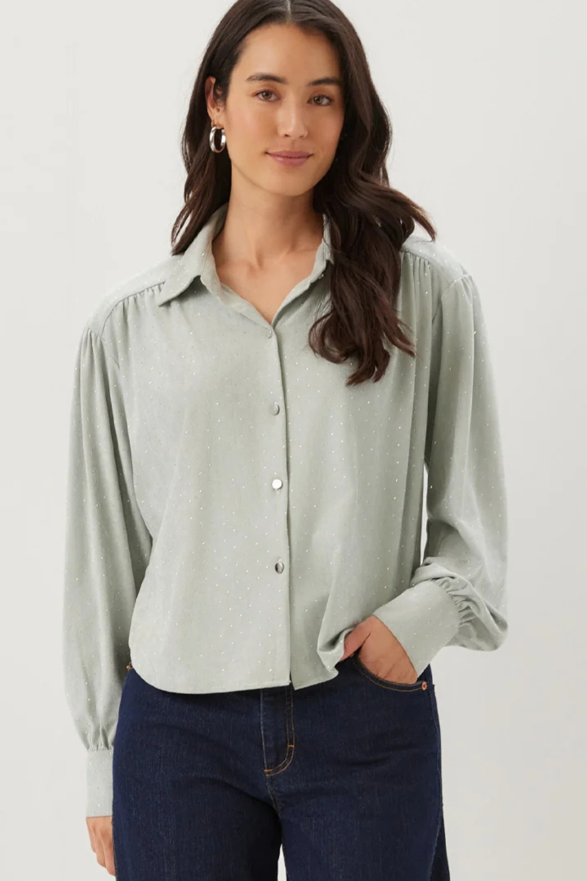A woman wearing a light sage green long-sleeve button-up blouse with gathered shoulders and dark blue jeans, standing against a plain light gray studio background.