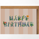 A square birthday card with a kraft envelope, featuring the words 'HAPPY BIRTHDAY' in blue letters decorated with small flowers and green leaves, set against a beige and pink striped background.