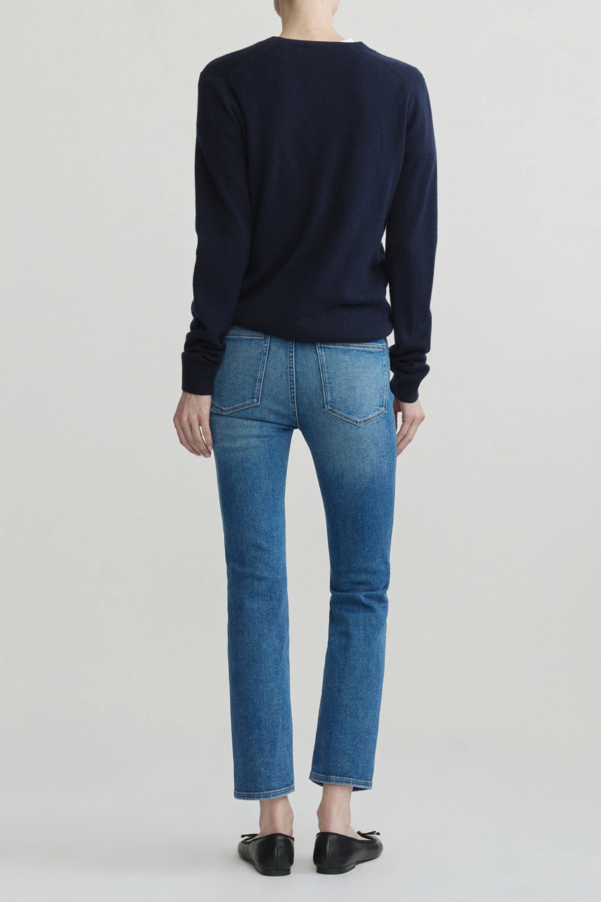 A model shown from the back wearing a dark navy long-sleeve crewneck sweater, blue cropped jeans, and black flats, set against a plain light gray studio background.