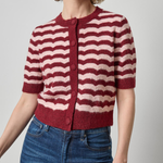 A woman wearing a short-sleeve knit cardigan with horizontal wavy stripes in burgundy and cream, paired with blue jeans, against a plain light gray studio background.