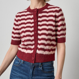 A woman wearing a short-sleeve knit cardigan with horizontal wavy stripes in burgundy and cream, paired with blue jeans, against a plain light gray studio background.