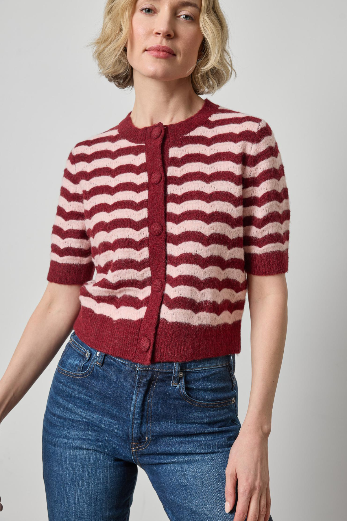 A woman wearing a short-sleeve knit cardigan with horizontal wavy stripes in burgundy and cream, paired with blue jeans, against a plain light gray studio background.