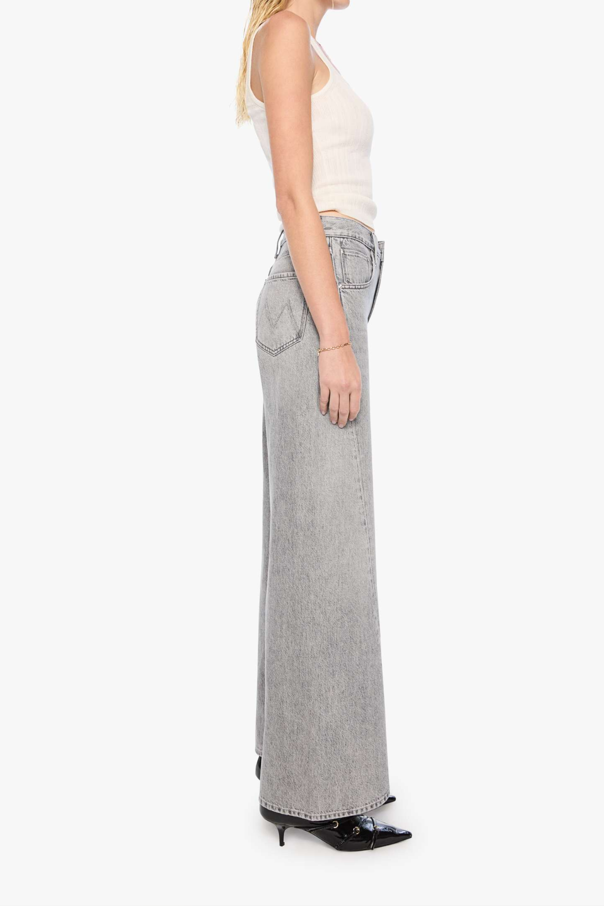 A side-profile view of a model wearing light gray wide-leg denim jeans, a cream ribbed tank top, and black pointed-toe heels against a plain white background.