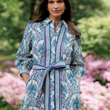 Woman wearing a blue patterned dress standing in a garden with pink flowers and green foliage.