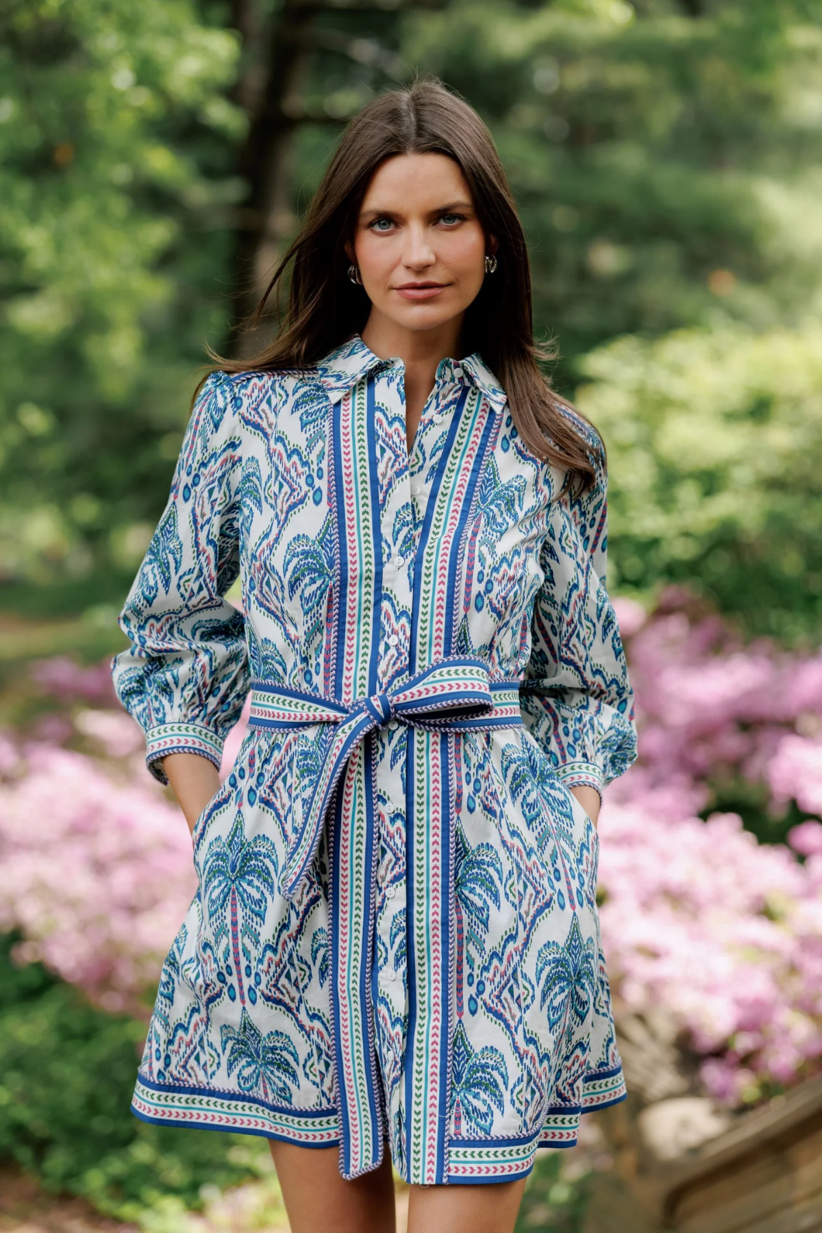 Woman wearing a blue patterned dress standing in a garden with pink flowers and green foliage.