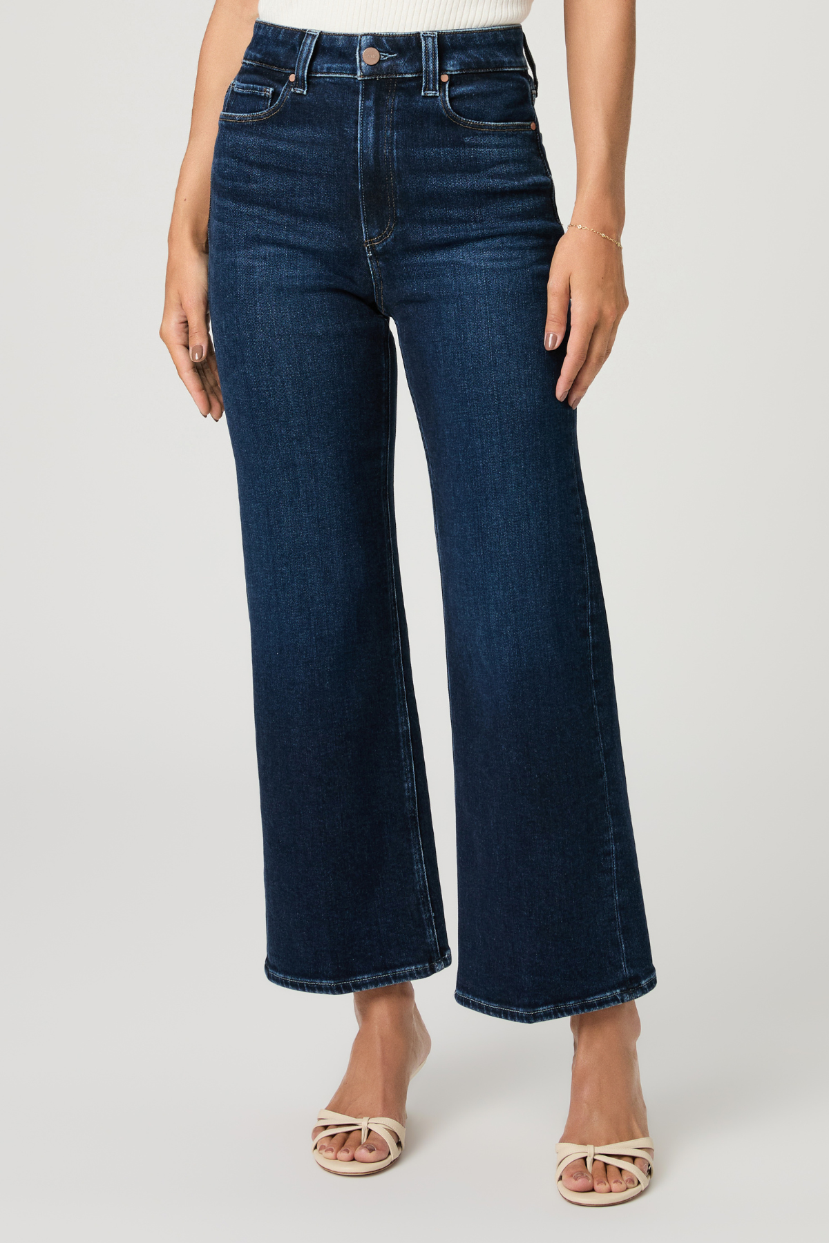 A model wearing dark blue high-waisted wide-leg cropped jeans with a faded wash, paired with beige heeled sandals, against a light gray studio background.