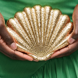 Gold shell-shaped clutch held by hands against a green fabric background