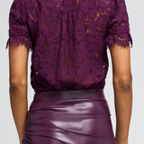 A woman shown from the back wearing a deep purple lace short-sleeve blouse and a matching purple faux-leather skirt against a light gray studio background.
