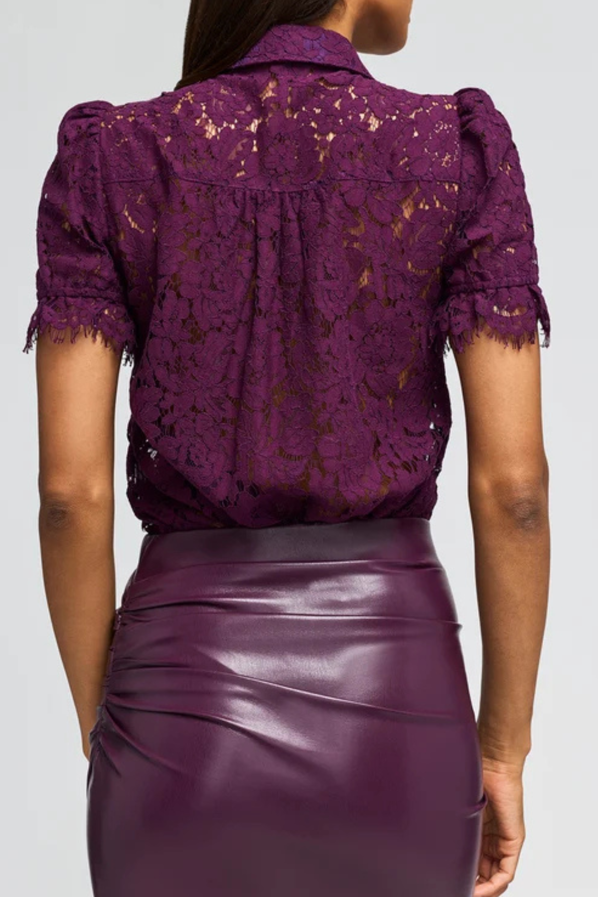 A woman shown from the back wearing a deep purple lace short-sleeve blouse and a matching purple faux-leather skirt against a light gray studio background.