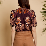 A woman viewed from behind wearing a short-sleeve blouse with a dark purple and orange floral pattern, paired with brown pants, set against a warm beige background with palm leaves.