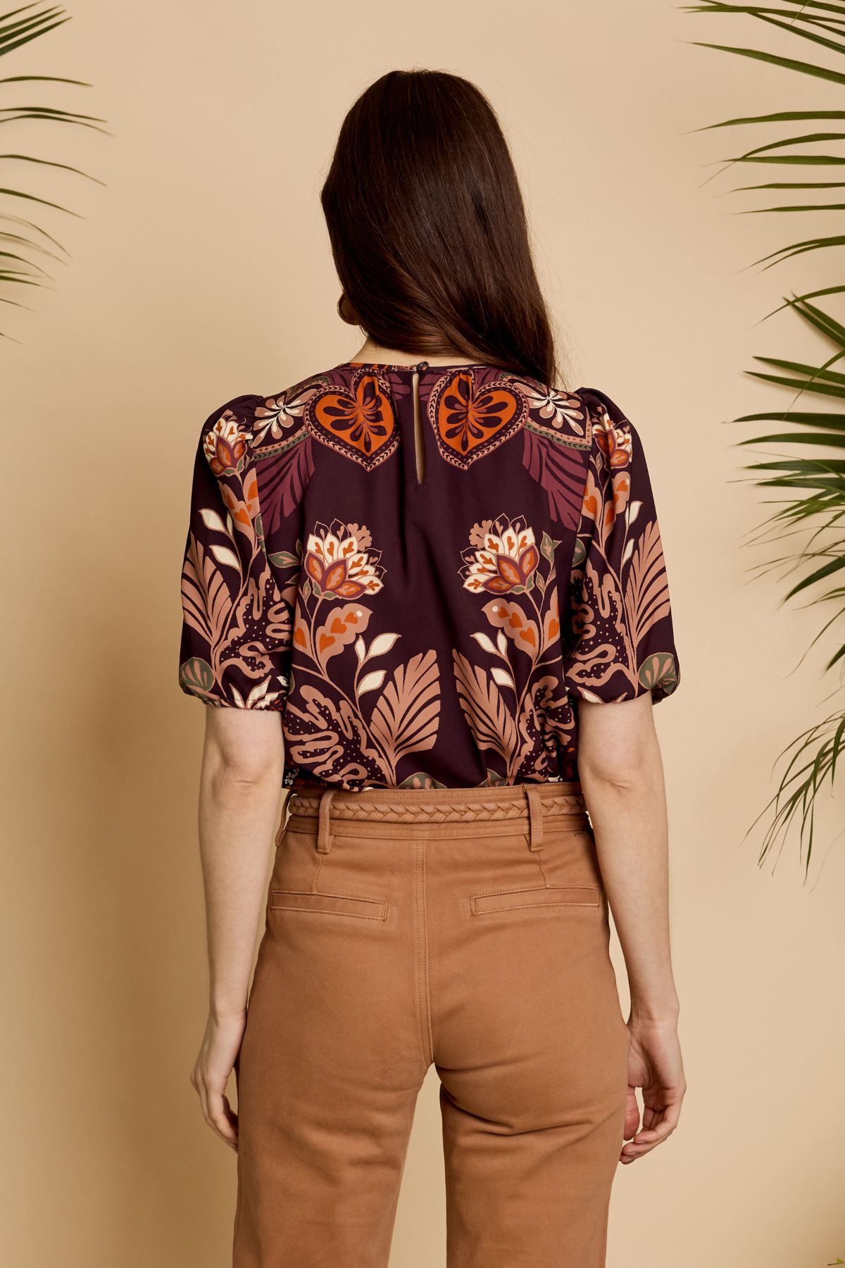 A woman viewed from behind wearing a short-sleeve blouse with a dark purple and orange floral pattern, paired with brown pants, set against a warm beige background with palm leaves.