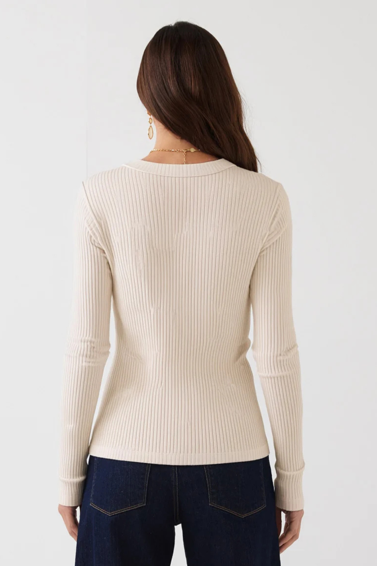 A woman viewed from the back wearing a cream ribbed long-sleeve sweater and dark blue jeans against a plain white background.