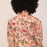 A person shown from the back wearing a light peach-pink button-up shirt with a colorful floral and bird print, featuring three-quarter ruffled sleeves and dark brown trousers, against a plain off-white background.