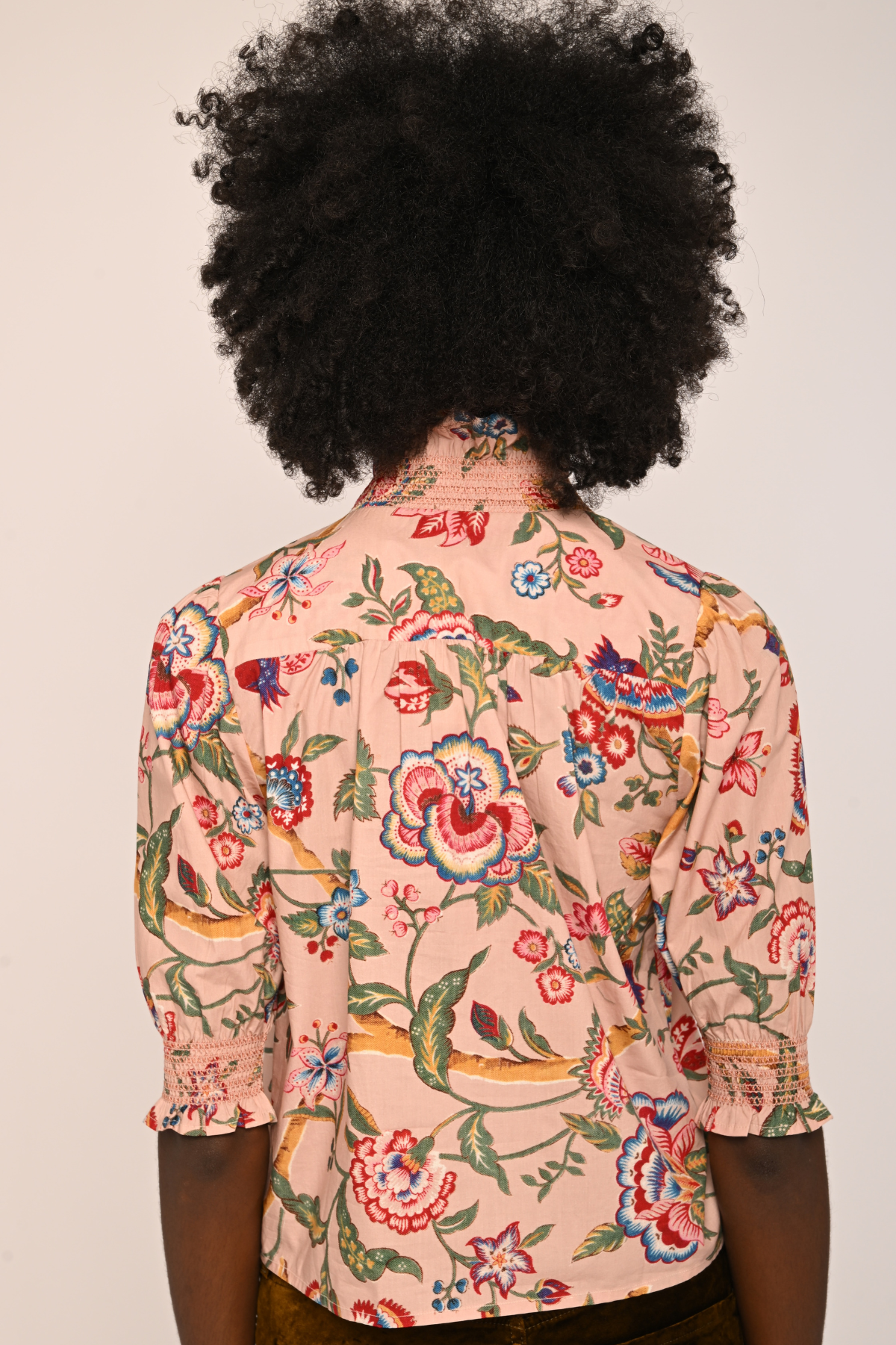 A person shown from the back wearing a light peach-pink button-up shirt with a colorful floral and bird print, featuring three-quarter ruffled sleeves and dark brown trousers, against a plain off-white background.