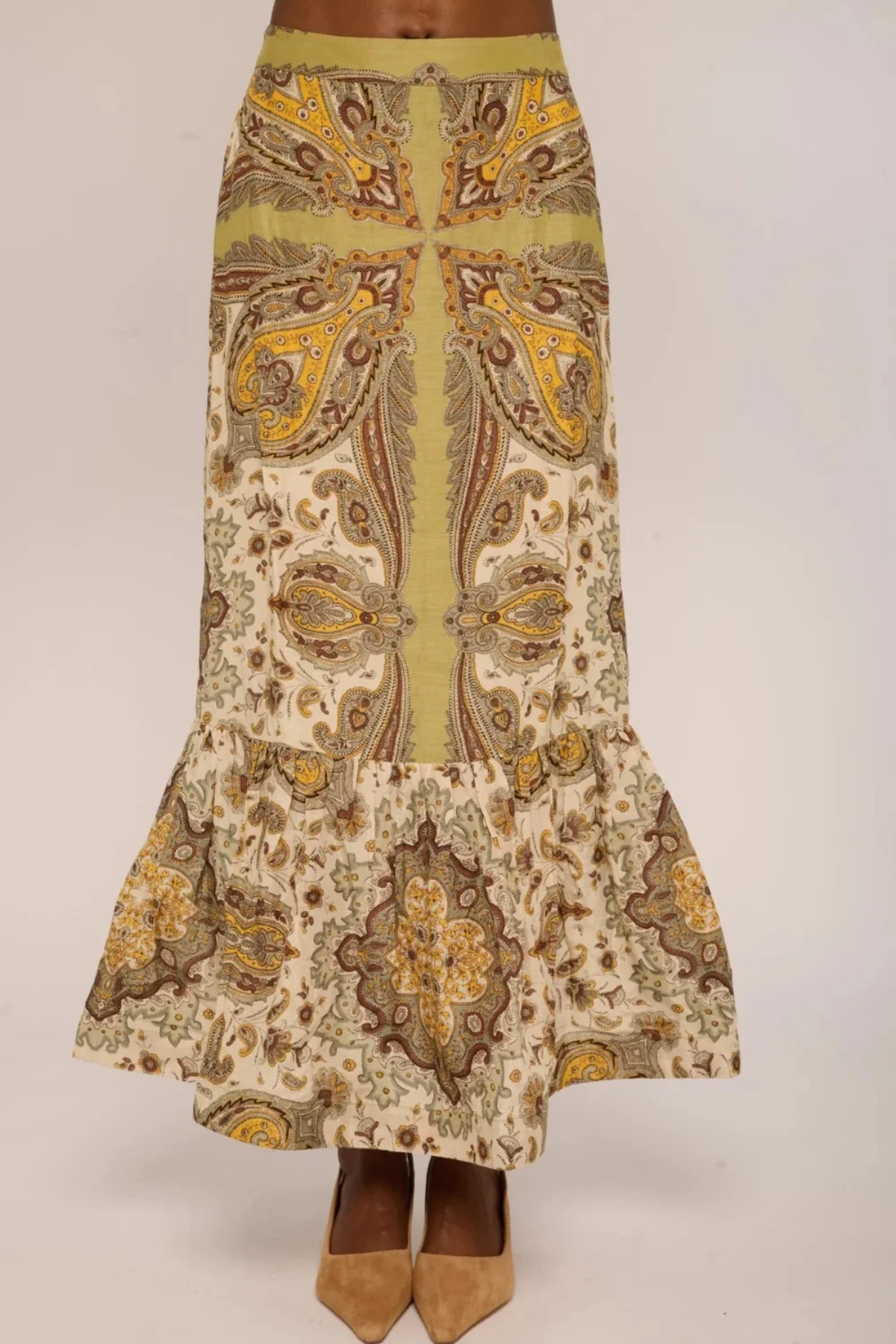 A long skirt with a ruffled hem, featuring a vertical olive green stripe down the center flanked by symmetrical paisley patterns in brown, gold, and beige. The skirt is shown on a model wearing tan pointed-toe heels against a plain light gray background.