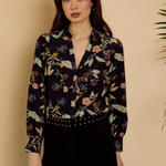 A woman modeling a long-sleeve button-up blouse with a dark navy floral and botanical print, paired with black pants, set against a warm beige background.