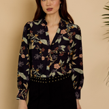 A woman modeling a long-sleeve button-up blouse with a dark navy floral and botanical print, paired with black pants, set against a warm beige background.
