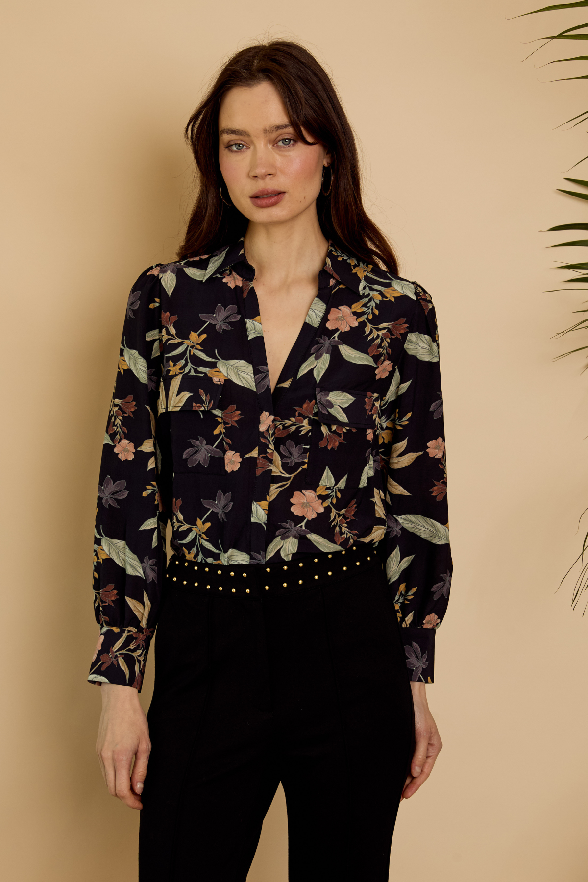 A woman modeling a long-sleeve button-up blouse with a dark navy floral and botanical print, paired with black pants, set against a warm beige background.