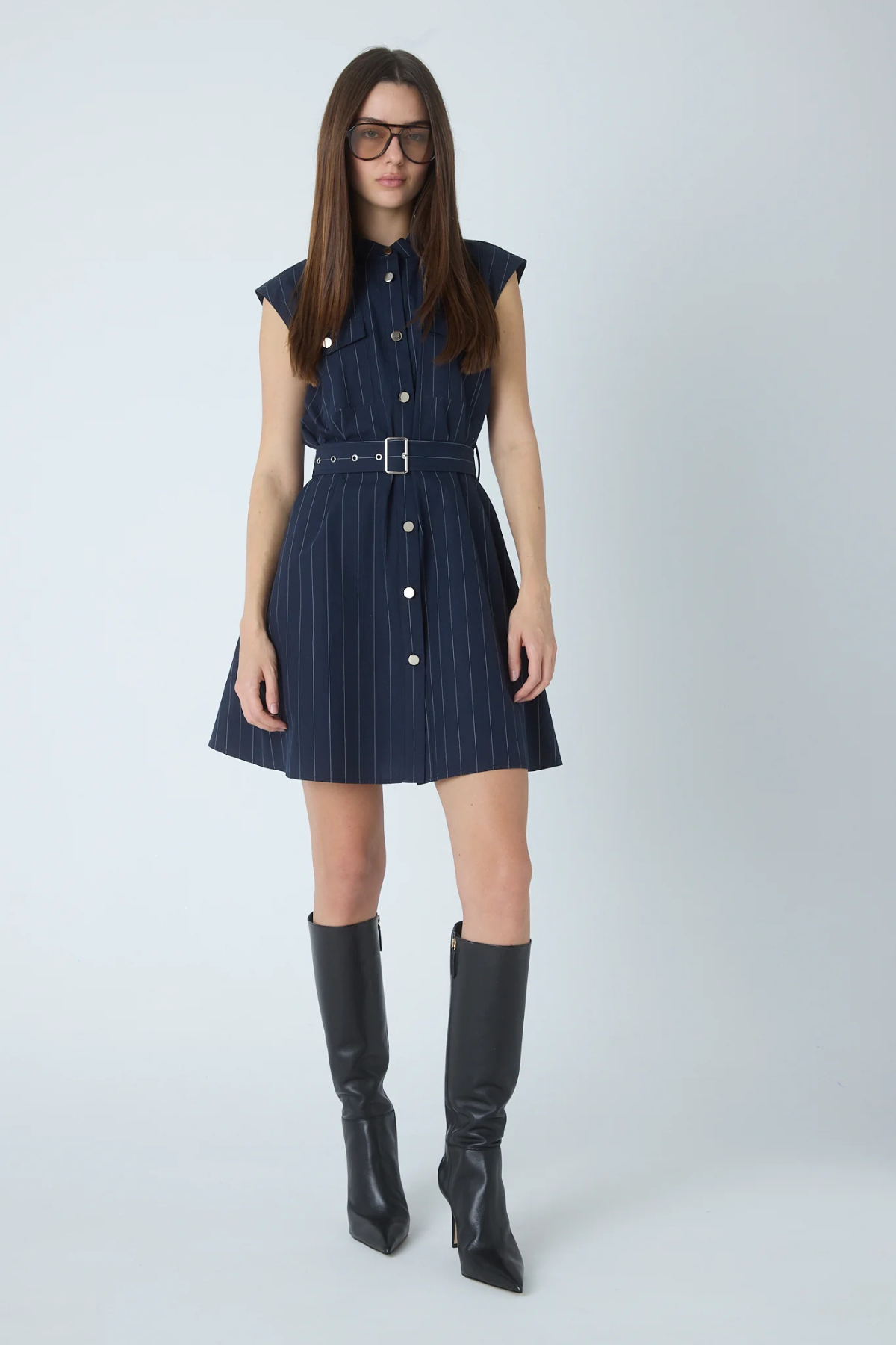 A woman modeling a sleeveless navy blue pinstriped mini dress with a matching belt and silver buckle, paired with black knee-high boots against a light gray studio background.