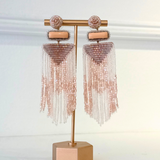 A pair of rose gold beaded tassel earrings displayed on a matching rose gold stand against a white background.