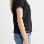 A woman shown from the side wearing a black short-sleeve crop top with white contrast stitching and blue jeans, against a plain light gray background.