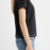 A woman shown from the side wearing a black short-sleeve crop top with white contrast stitching and blue jeans, against a plain light gray background.