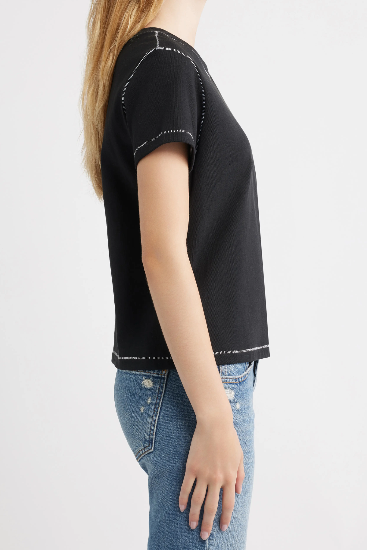 A woman shown from the side wearing a black short-sleeve crop top with white contrast stitching and blue jeans, against a plain light gray background.