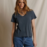 A woman with long light brown hair wearing a dark slate blue-gray v-neck t-shirt and blue jeans, standing against a neutral beige fabric backdrop.