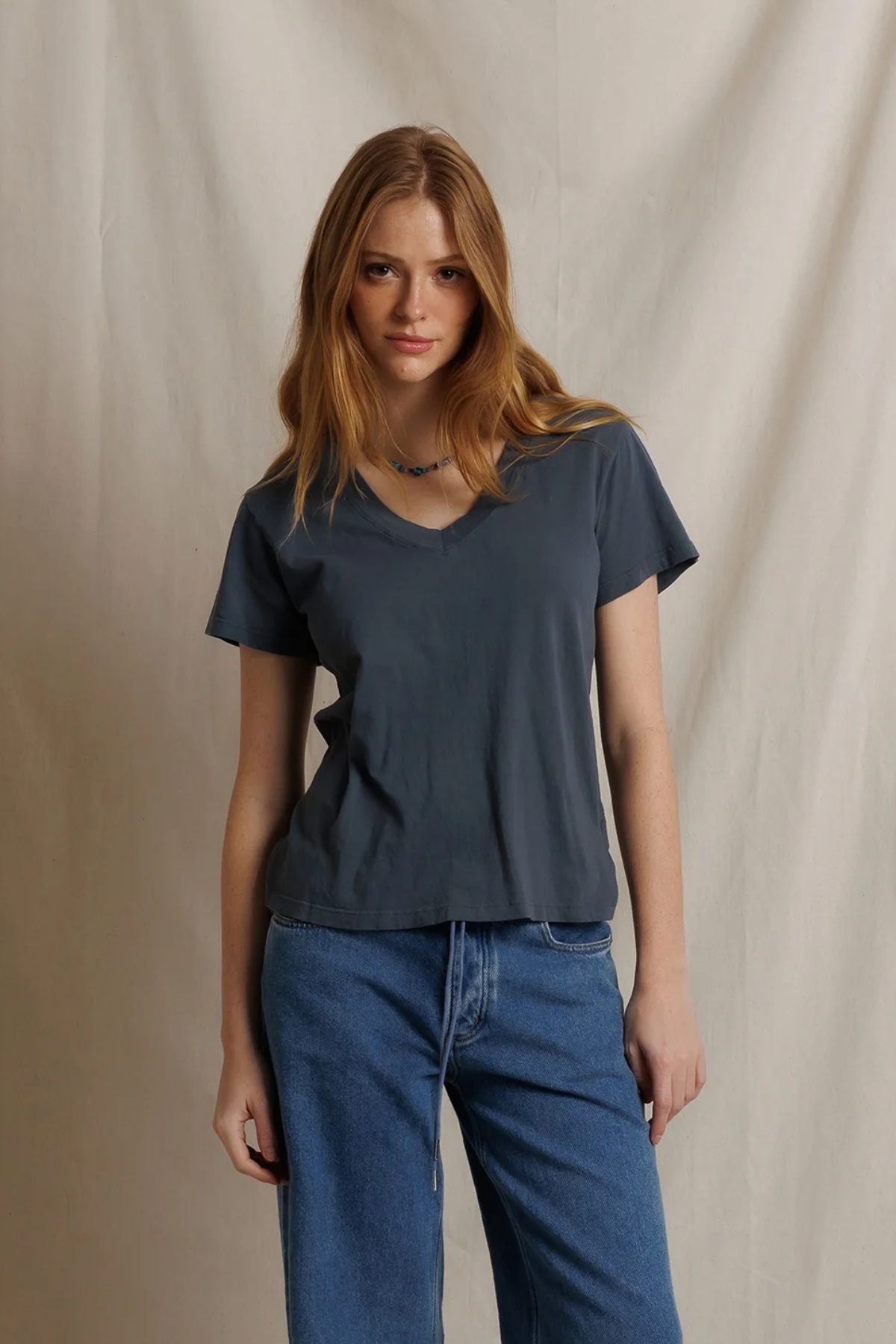 A woman with long light brown hair wearing a dark slate blue-gray v-neck t-shirt and blue jeans, standing against a neutral beige fabric backdrop.