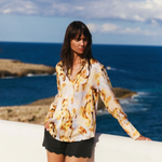 Woman in a patterned top and black shorts standing by the ocean with a clear blue sky.