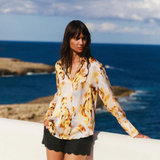 Woman in a patterned top and black shorts standing by the ocean with a clear blue sky.
