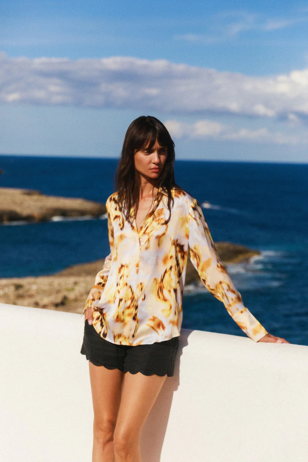 Woman in a patterned top and black shorts standing by the ocean with a clear blue sky.