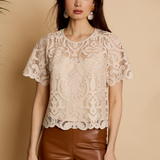 A woman modeling a short-sleeve cream lace blouse with a round neckline and brown faux-leather pants, standing against a warm beige background.