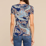 A woman viewed from the back wearing a fitted short-sleeve top with an ornate paisley-style print in navy, light blue, beige, and rust tones, paired with blue jeans against a plain beige studio background.